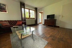 A living room with a glass coffee table, a brown leather sofa, a wooden cabinet with a television, and a large window with curtains. - Holiday apartment with 2 bedrooms (Schleiden-Broich)