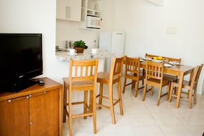 A compact kitchen with a TV, wooden chairs, a dining table, and a refrigerator. - Pousada Bora Bora (Bombinhas)