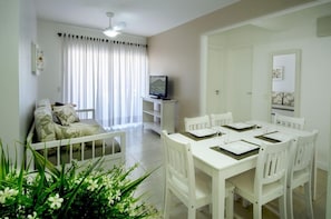 Una habitación con una mesa de comedor lista para cuatro personas, un televisor en un mueble bajo, un sofá con cojines y una ventana con cortinas.