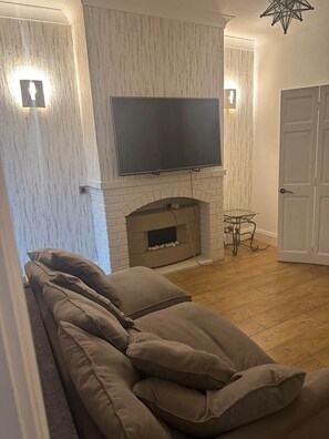 En stue med sofa, pejs, tv ophængt over pejsen, og en dør til højre.