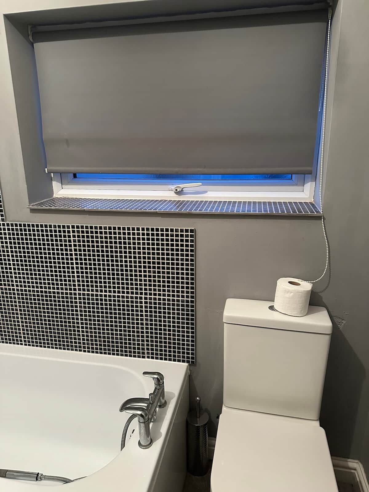 Ett badrum med badkar, toalett och ett fönster som släpper in blått ljus.