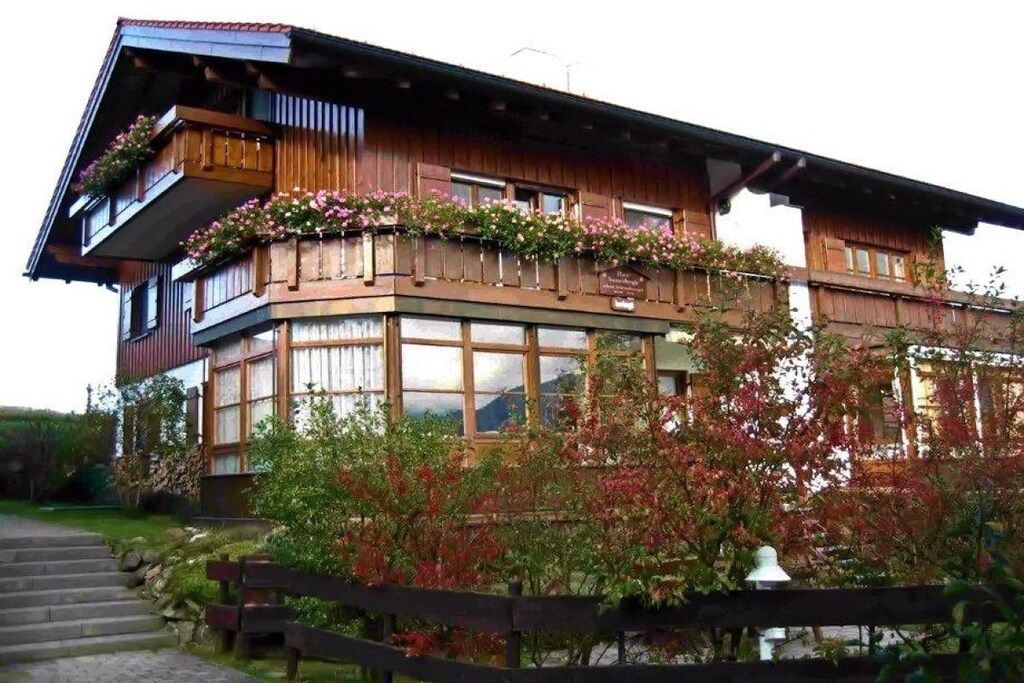 Un chalet in legno con un balcone decorato da fiori, ampie finestre e una scala in pietra che conduce all'ingresso.