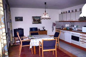 Uma sala de jantar com mesa posta para quatro pessoas, televisão e área de cozinha com armários e eletrodomésticos.