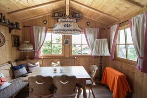 Interior de una cabaña de madera con una mesa de comedor, sillas, un banco y una ventana con cortinas.