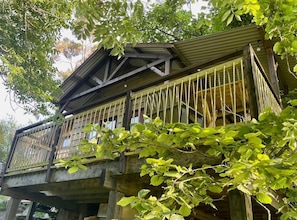 A wooden structure with a balcony, supported by pillars, surrounded by foliage.