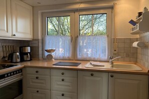 A kitchen with a stove, coffee maker, and a window with curtains. - Holiday home with 1 bedroom (Varel - Dangast)