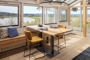 A modern dining area with a wooden table and chairs, large windows with a view of a river and landscape, and a built-in bench with cushions. - Holiday apartment in Kronburg with sauna (Kronburg)