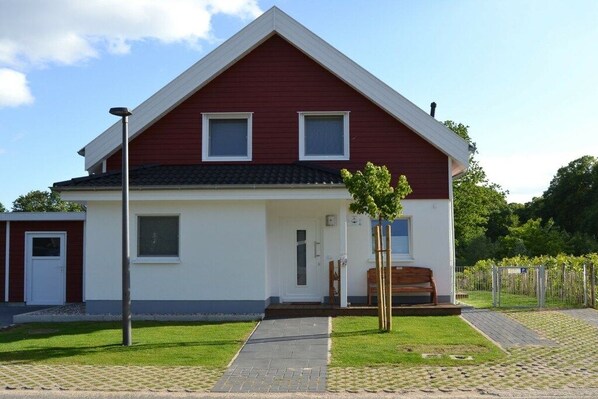 Uma casa de dois andares com fachada vermelha e detalhes brancos, varanda frontal com um banco e uma pequena árvore no jardim da frente.