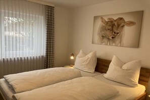 Cama de casal com roupa de cama branca e cabeceira, abajur e quadro de duas vacas fixado na parede.