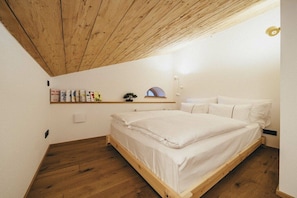 A bedroom with a wooden ceiling, a bed with white bedding, a shelf with books and a small plant, and a white wall. - high-quality with sauna (Ruhpolding)