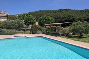 A swimming pool with a clear blue water surface, surrounded by a grassy area with trees and a wooden pergola with lounge chairs. - Bastide des Grandes Terres gite Alpilles (MERINDOL)