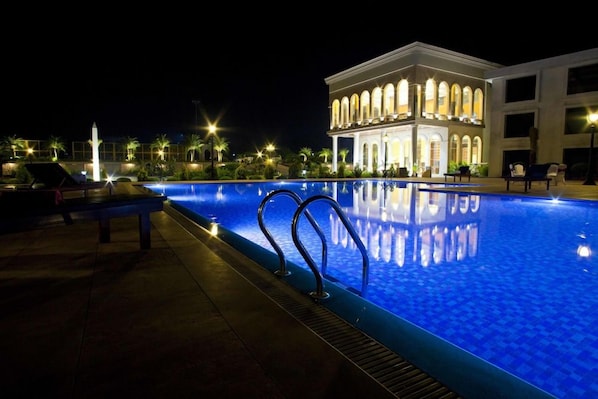 Ein beleuchteter Pool mit einer Fontäne, umgeben von Palmen und einem Gebäude mit Bogenfenstern.