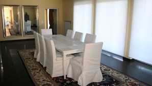 A dining area with a white table and chairs, a patterned rug, and large windows with blinds.