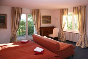 A hotel room with a bed, a sofa, a desk, and a chair. There is a window with curtains and a view of greenery outside. - "Stutengarten Estate" (Öhningen)
