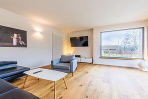 Ein modernes Wohnzimmer mit Sofa, Sessel, Couchtisch und einem an der Wand befestigten Fernseher.