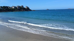 A beach with waves, a clear sky, and buildings in the background. - Dinard-ville location 2 chambres, plage de l'écluse-commerces-transports à pieds (Dinard)