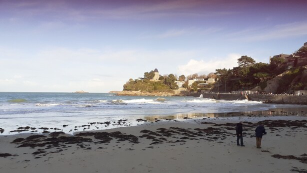 A beach with waves, a person standing, and buildings in the background. - Dinard-ville location 2 chambres, plage de l'écluse-commerces-transports à pieds (Dinard)