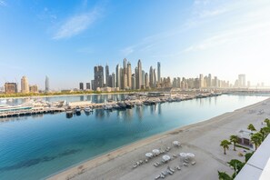A marina with boats docked, a sandy beach with umbrellas, and a city skyline in the background. - StayShort - Stunning Beachfront 3-Bedroom Apartment Sleeps 5 with Infinity Pool, Skyline Views & Private Beach Access (Emaar Beach Front)