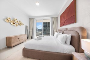 A modern hotel room with a large bed, a wooden dresser, and a city view. - StayShort - Stunning Beachfront 3-Bedroom Apartment Sleeps 5 with Infinity Pool, Skyline Views & Private Beach Access (Emaar Beach Front)