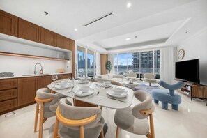 A modern apartment with a dining area, kitchen, living room, and a large window with a city view. - StayShort - Stunning Beachfront 3-Bedroom Apartment Sleeps 5 with Infinity Pool, Skyline Views & Private Beach Access (Emaar Beach Front)