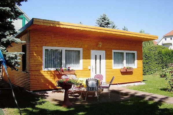 Ein kleines Holzhaus mit gelbem Anstrich, weißen Fenstern und einer Tür. Vor der Veranda stehen zwei Kübelpflanzen und ein Tisch mit Stühlen.
