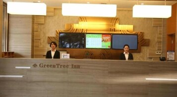Un mostrador de recepción en GreenTree Inn con dos recepcionistas, una pantalla digital y un área de recepción de madera.