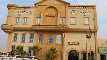 A building with multiple signs in Arabic, including 'Al-Sabah Hotel' and 'Al-Sabah Hotel'. There are flags, palm trees, and parked cars in front.