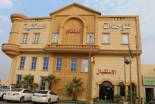 Een gebouw met meerdere borden in Arabisch, waaronder 'Al-Sabah Hotel' en 'Al-Sabah Hotel'. Voorop zijn vlaggen, palmbomen en geparkeerde auto's te zien.