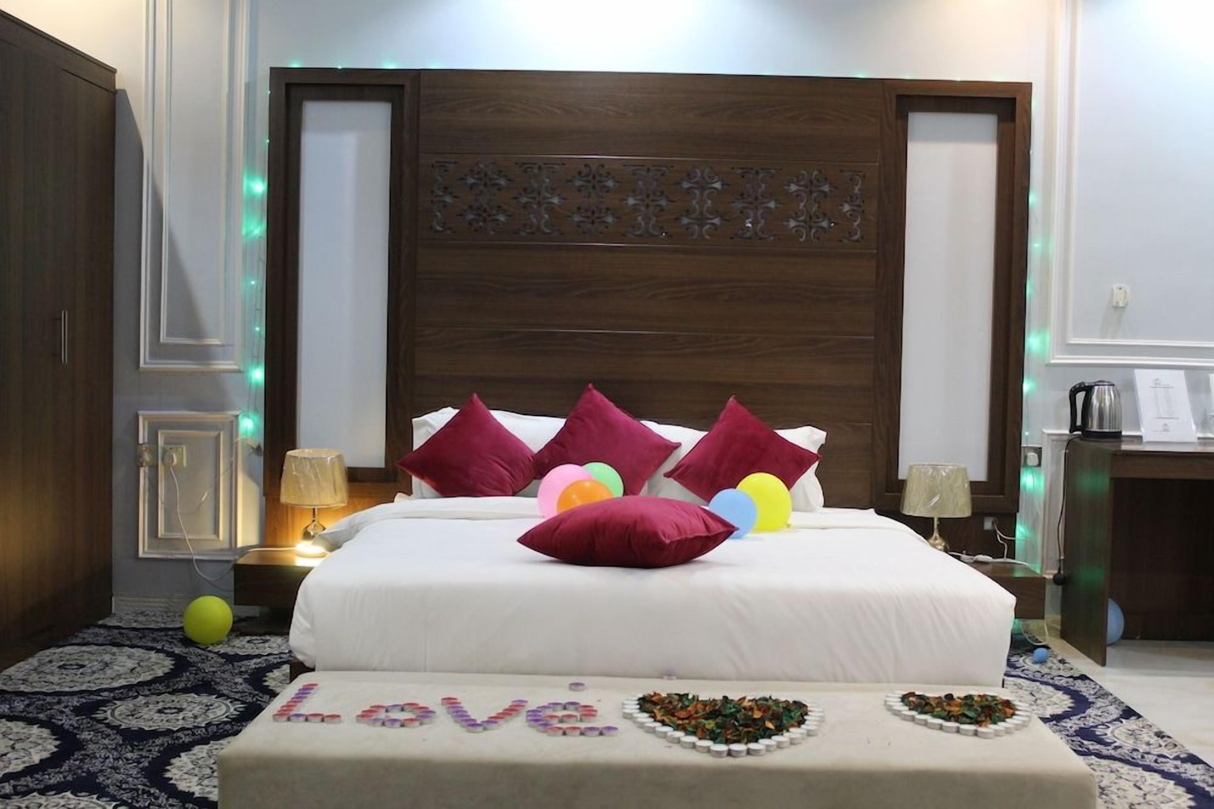 A hotel room with a large bed, multiple pillows, a bedside table with a lamp, and a decorative wall panel.