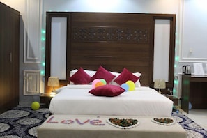 Een hotelkamer met een groot bed, meerdere kussens, een nachtkastje met een lamp en een decoratief wandpaneel.