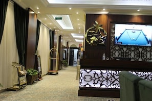 Een hotel lobby met een receptiebalie, een televisiescherm waarop een tafelvoetbalspel te zien is, en een gang die naar andere ruimtes leidt.
