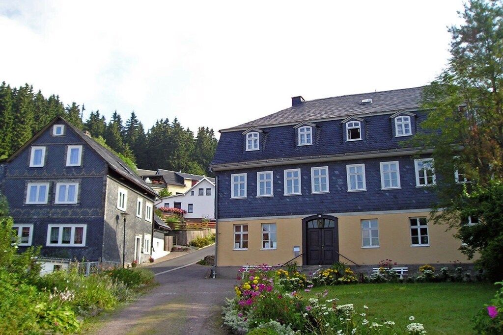 A two-story building with a dark roof and light walls, a driveway leading to the entrance, and a garden with various flowers.