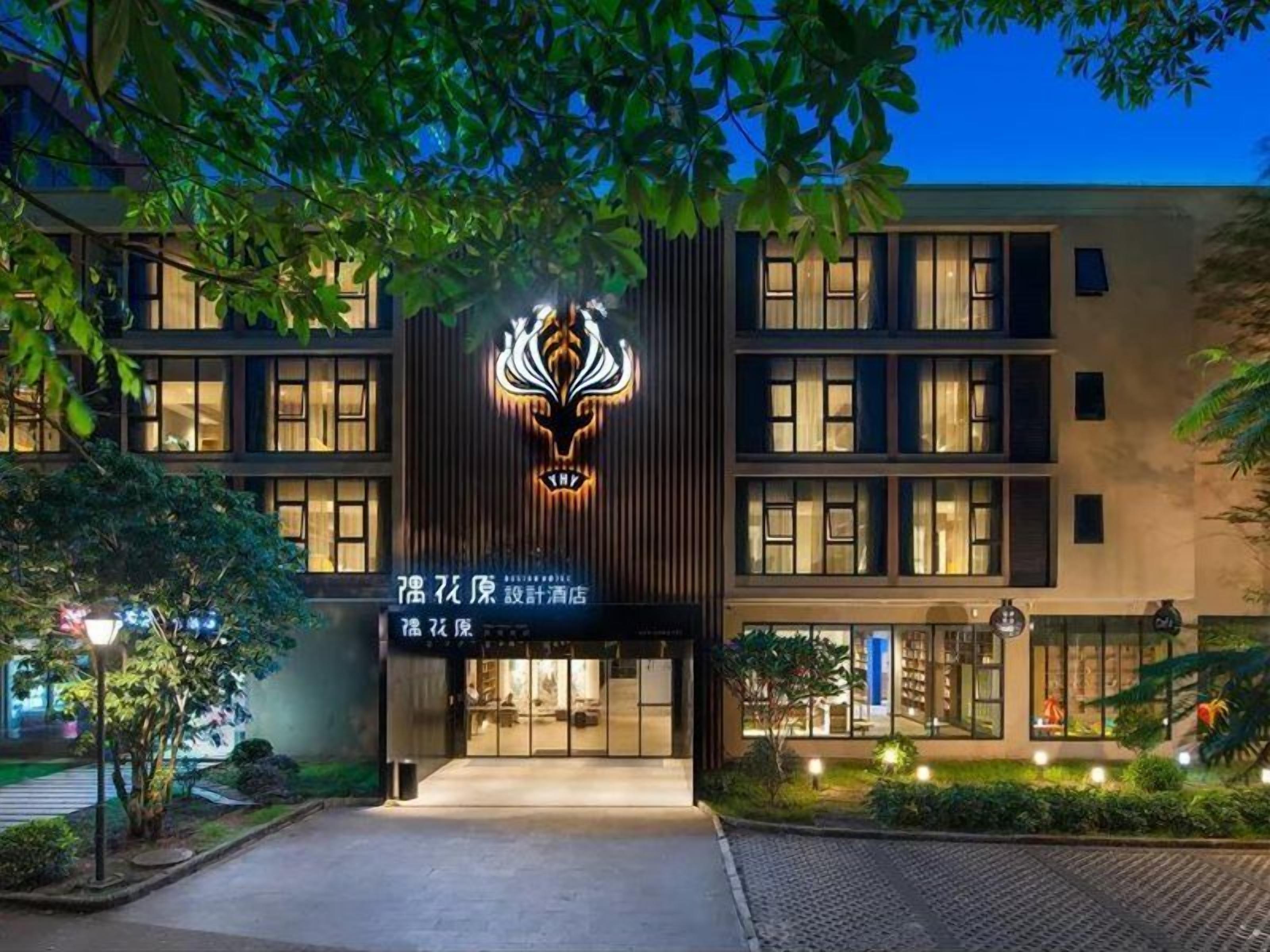 A modern building with a prominent illuminated logo at the entrance, a well-lit facade, and a landscaped area with trees and shrubs.