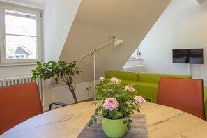 Ein Zimmer mit einem Esstisch, einer Topfpflanze mit Blumen, einem grünen Sofa und einem an der Wand befestigten Fernseher.