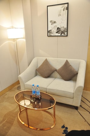 Un salon moderne avec un canapé, deux coussins, une table basse en verre sur laquelle reposent des bouteilles d’eau, et une œuvre d’art encadrée accrochée au mur.