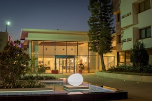 Entrada de hotel con fachada de vidrio, una recepción y una zona de piscina con una estructura esférica distintiva.