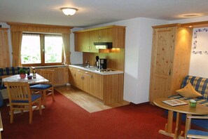 Cozinha compacta com armários de madeira, espaço de refeições com mesa e cadeiras, e área de estar pequena com sofá e mesa lateral.
