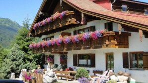 Een klassiek chalet met houten balkons, versierd met bloemen, een stenen bankje en een berg op de achtergrond.
