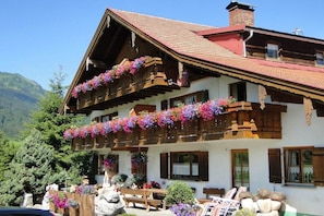 Um chalé tradicional com varandas de madeira decoradas com flores, um banco de pedra e montanhas ao fundo.