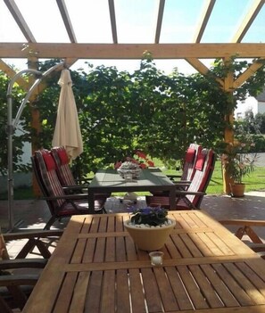Een terras buiten met een houten tafel en stoelen, een hangende parasol en uitzicht op een tuin.
