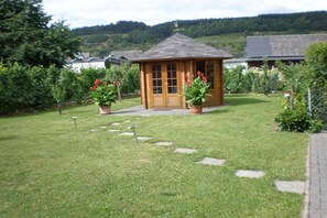Eine kleine Holzhütte mit Tür und Fenstern, umgeben von einem gepflegten Rasen und Topfpflanzen.