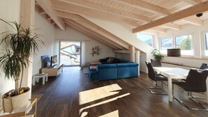 A spacious loft with a high ceiling, a dining area with a table and chairs, a blue sofa, and a large window providing a view of the outdoors. - "Penthouse" Alpine Bliss (Füssen)