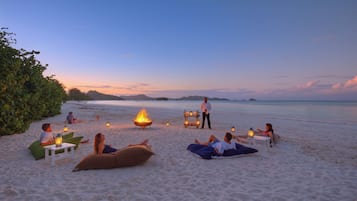 Escena de playa con gente relajándose en esterillas y sillas, un brasero y una persona sosteniendo un farol.