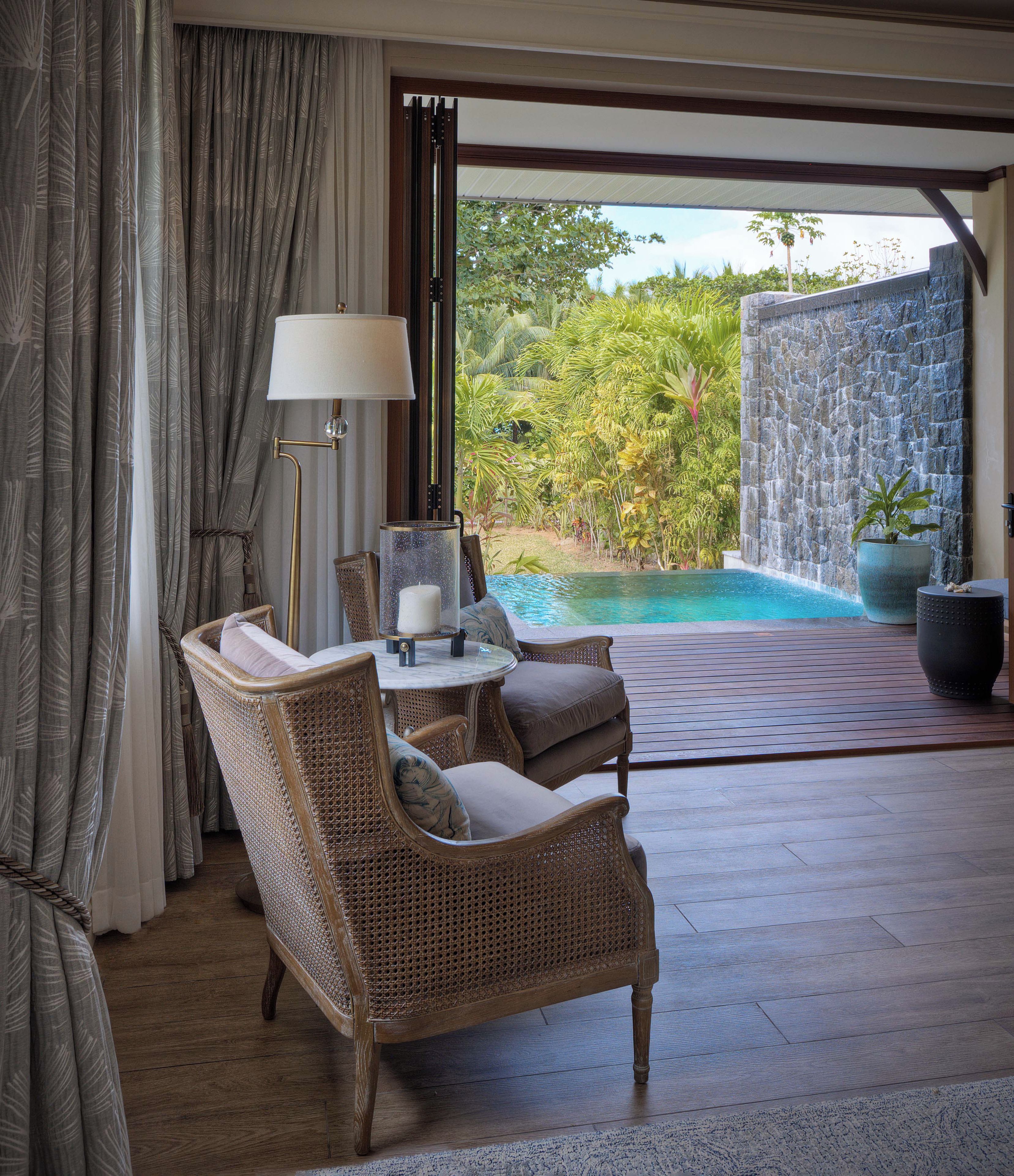 Quarto com mobiliário de vime, uma mesa com abajur e vista para uma piscina e vegetação.