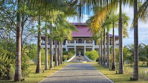 A building with a central entrance, flanked by palm trees, and a paved pathway leading to it. - La Cigale Estate (Anse Volbert Village)