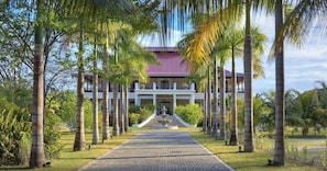 A building with a central entrance, flanked by palm trees, and a paved pathway leading to it. - La Cigale Estate (Anse Volbert Village)