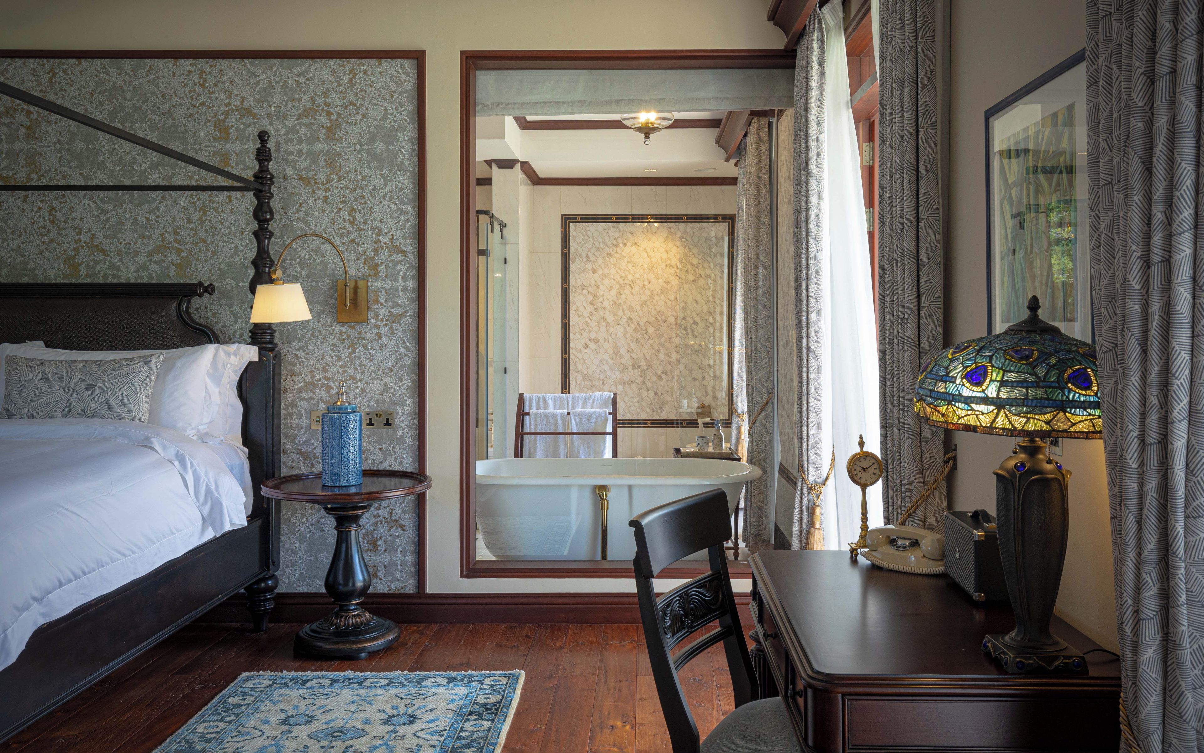 Habitación de hotel con cama, mesita de noche, lámpara, silla y vistas al baño.