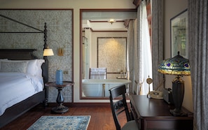 Habitación de hotel con cama, mesita de noche, lámpara, silla y vistas al baño.