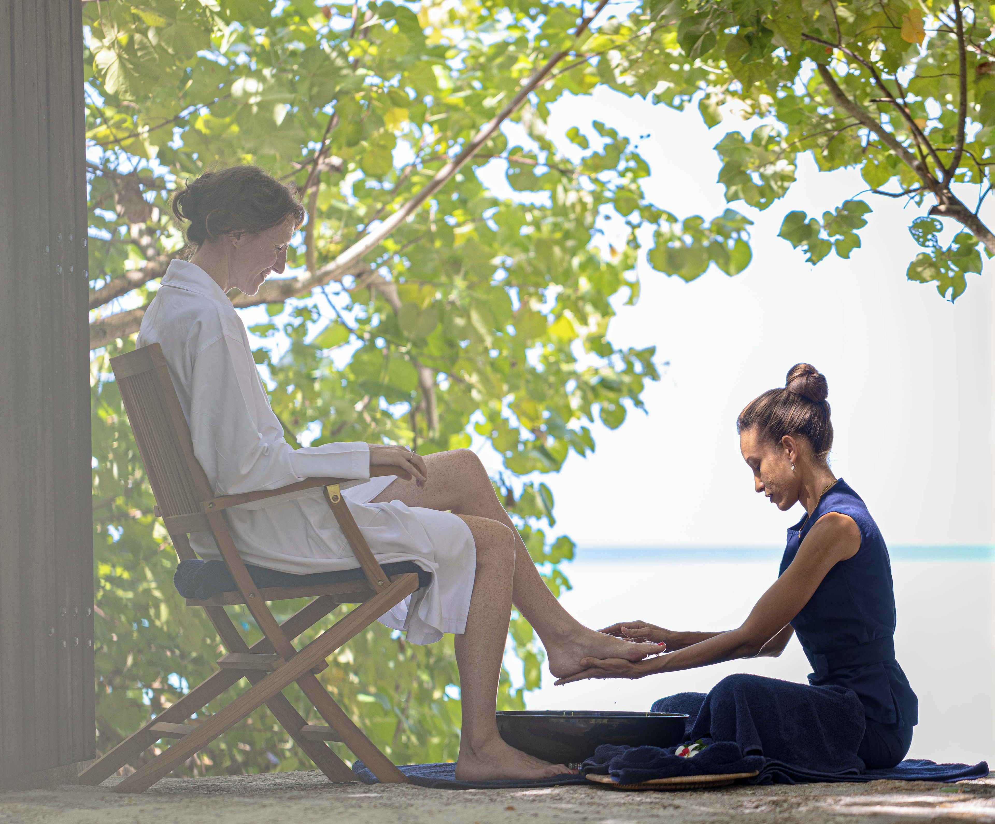 Two individuals seated on a wooden chair outdoors, one performing a foot massage on the other.