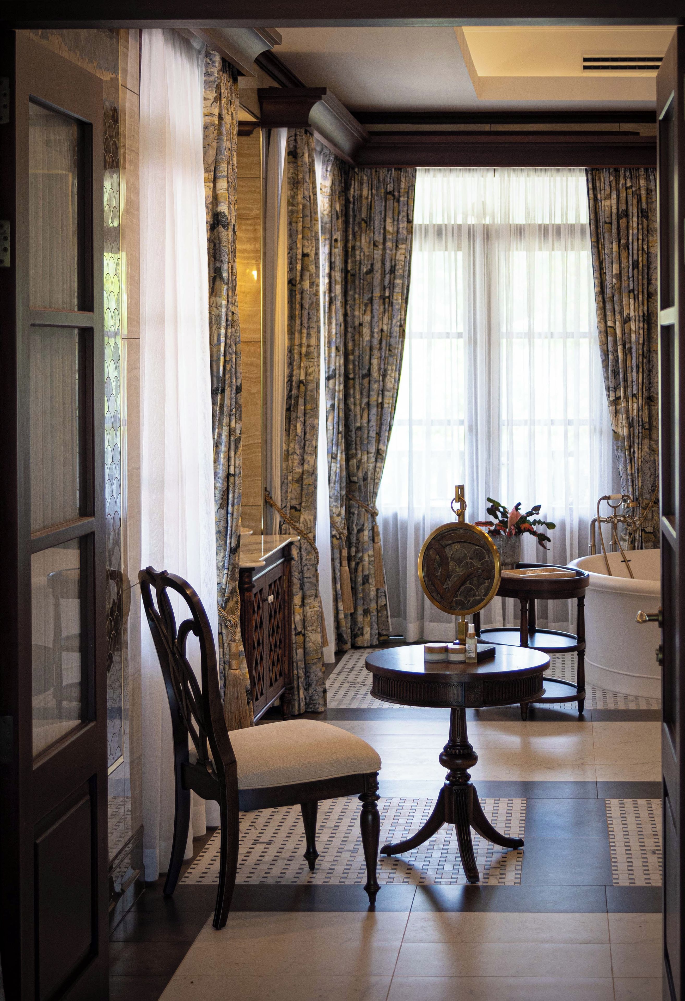 Una habitación con una silla de madera, una mesa redonda con un espejo y una bañera al fondo.